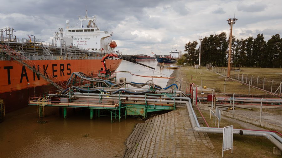 Un buque tanque rojo amarrado junto al muelle de una terminal, con caños y mangueras de carga sobre la plataforma y árboles en la orilla.