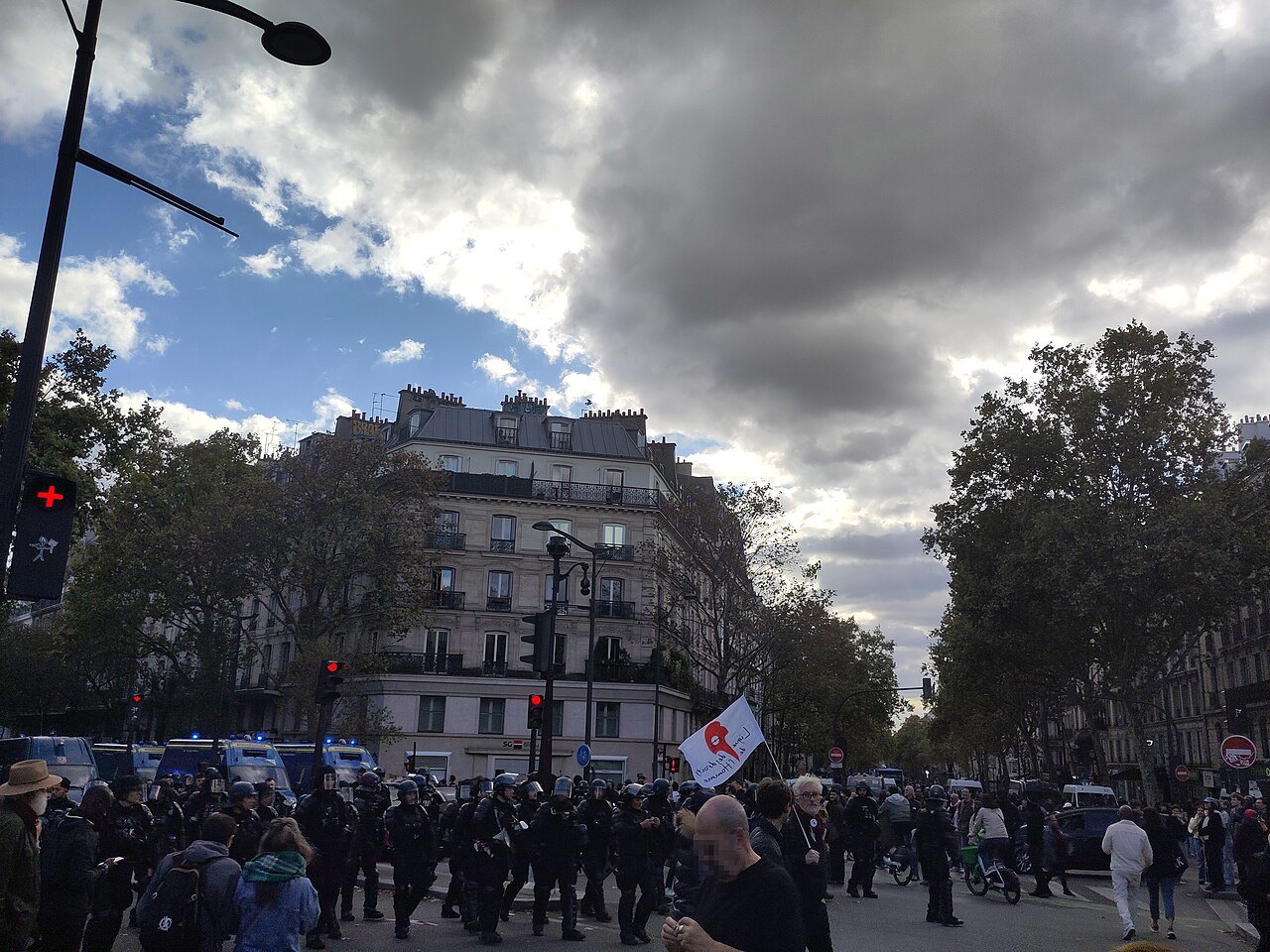 Una fila de policías antidisturbios con cascos camina por un bulevar de París entre manifestantes, con camionetas policiales detrás y una bandera de la Liga de los Derechos Humanos en alto