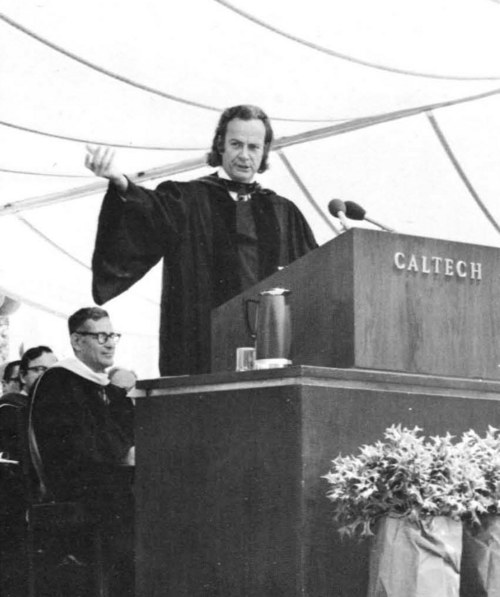 Richard Feynman, con toga académica, habla con el brazo extendido detrás de un atril con la palabra CALTECH, bajo una carpa