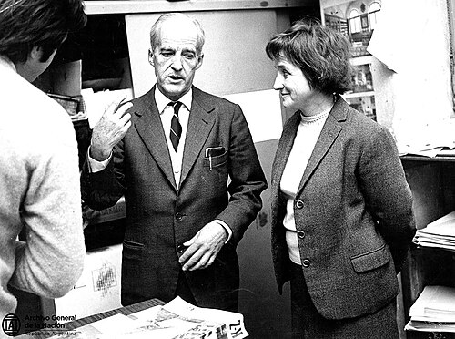 Luis F. Leloir, de traje y corbata, habla de pie en el instituto de la Fundación Campomar junto a una mujer que sonríe; en primer plano, de espaldas, una persona que lo escucha