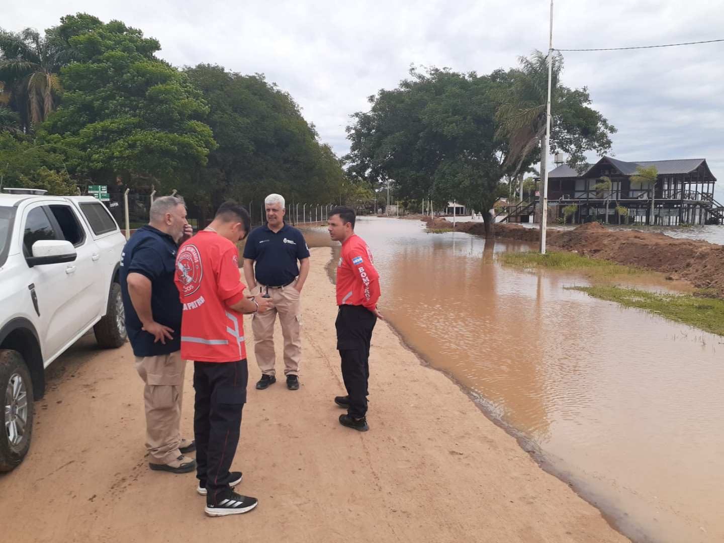 Cuatro hombres, dos de ellos con remeras rojas de bomberos voluntarios y dos con camisas azules, conversan junto a una camioneta blanca en un camino de tierra; a la derecha, la calle y los terrenos están cubiertos por agua marrón y hay un terraplén de tierra.
