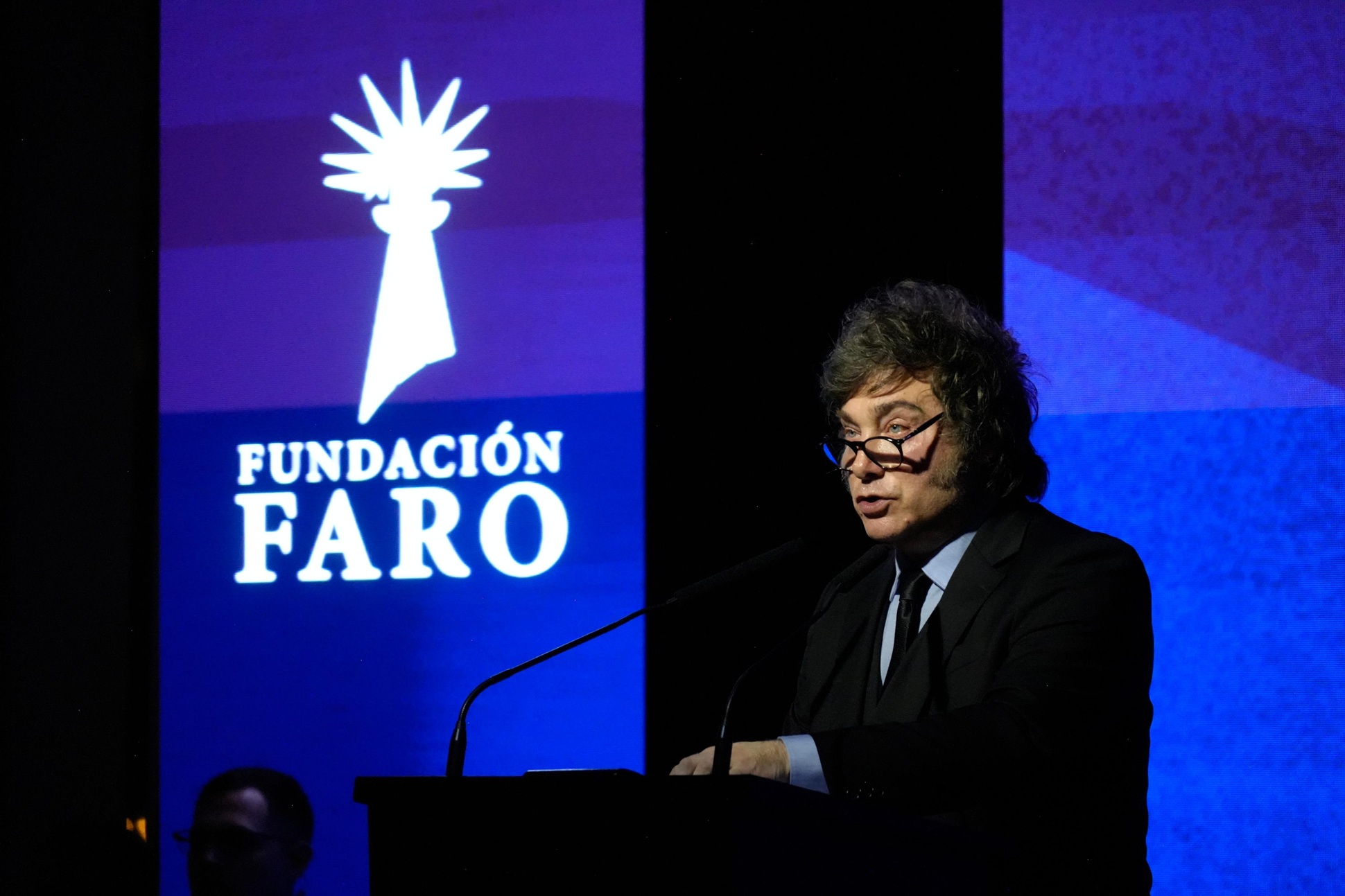 Javier Milei habla desde el atril en la cena de la Fundación Faro, con el logo de la fundación proyectado detrás.