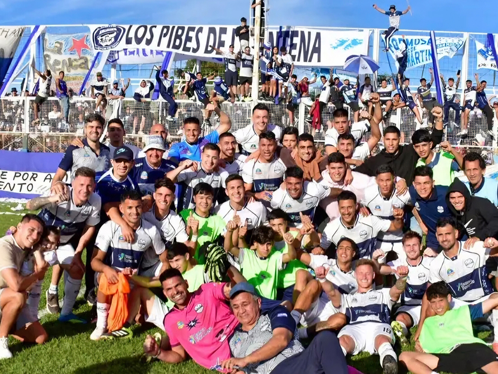 Jugadores y cuerpo técnico de San Martín de Formosa posan festejando sobre el césped, con hinchas y banderas detrás