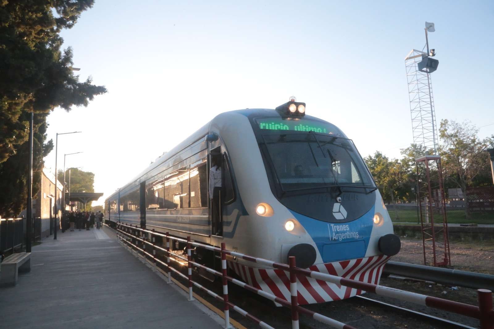 Un tren de pasajeros blanco y celeste de Trenes Argentinos, con franjas rojas y blancas en el frente, detenido junto al andén de una estación del Alto Valle, con pasajeros al fondo