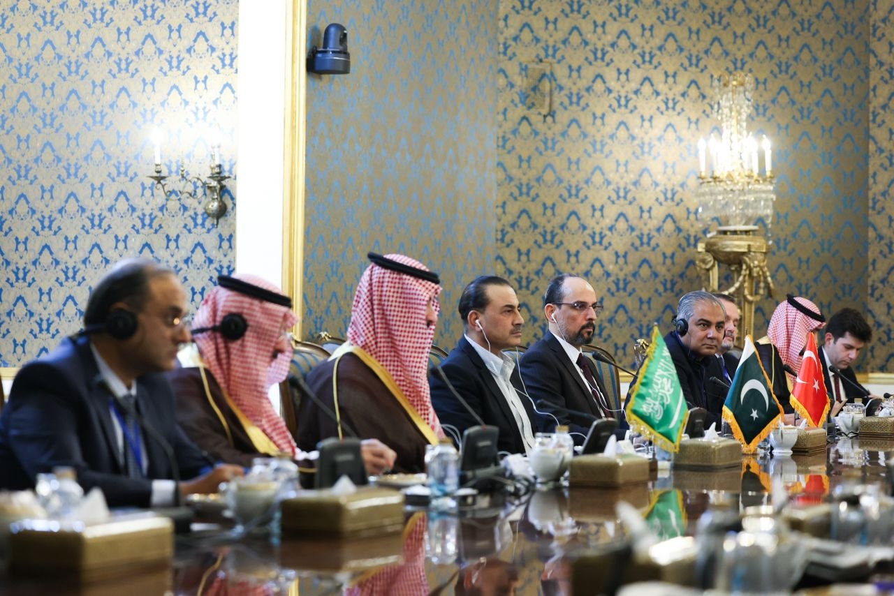 Mesa de reunión larga en un salón con arañas de cristal: de un lado, miembros de las delegaciones saudí, paquistaní y turca, con banderitas de Arabia Saudita, Pakistán y Turquía sobre la mesa.
