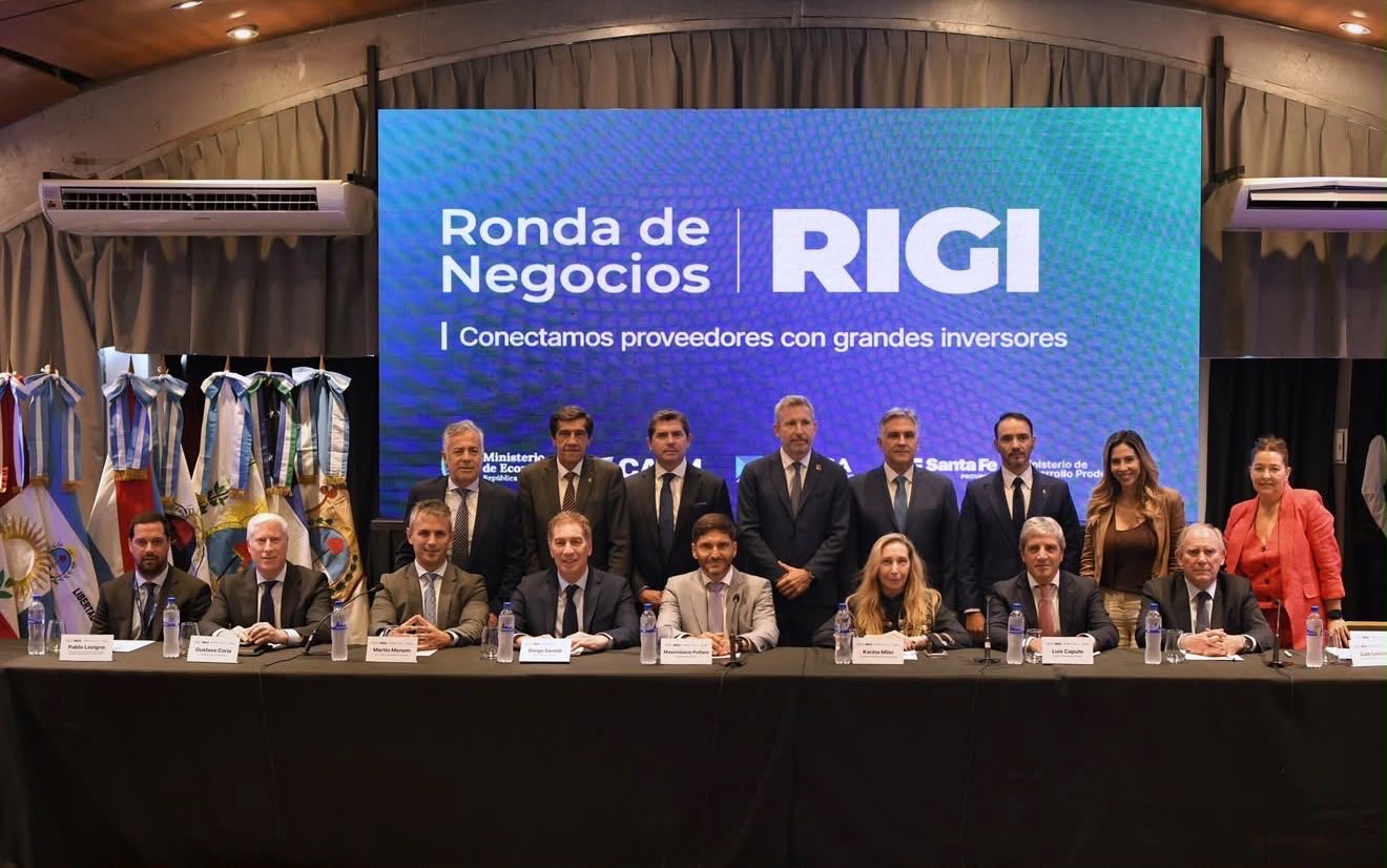 Luis Caputo, Karina Milei, Maximiliano Pullaro, Diego Santilli, Martín Menem y otros funcionarios y gobernadores, sentados a una mesa y de pie, frente a una pantalla que dice «Ronda de Negocios RIGI. Conectamos proveedores con grandes inversores»