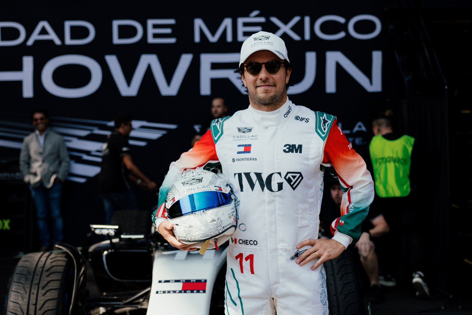 Sergio Pérez, con un buzo antiflama blanco con detalles verdes y rojos, sostiene su casco junto al auto antes de la exhibición en Ciudad de México.