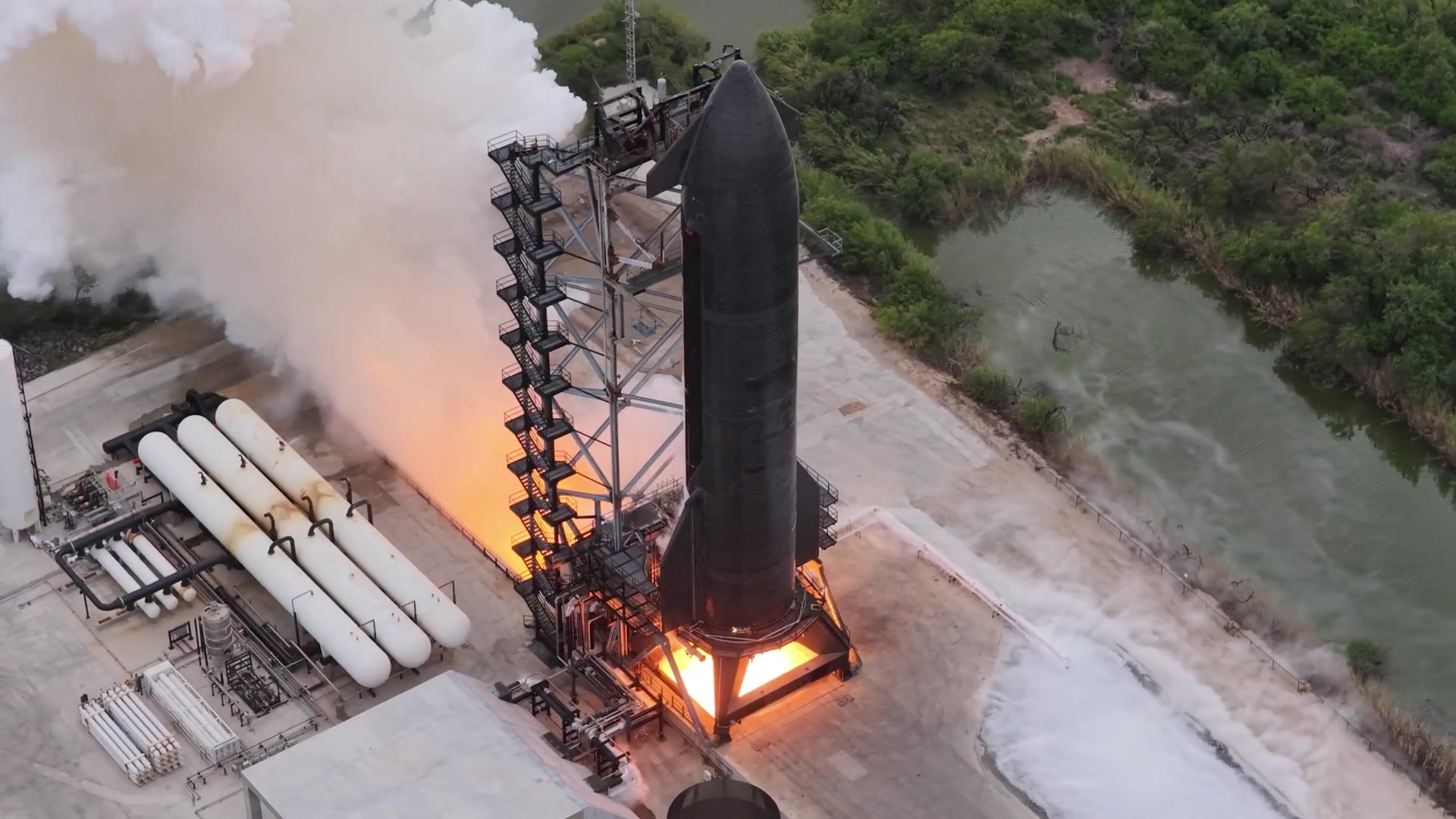 Vista aérea de la nave Starship Ship 42, negra, sujeta a un soporte de prueba junto a una torre metálica, con llamas en su base y una nube de vapor a un costado