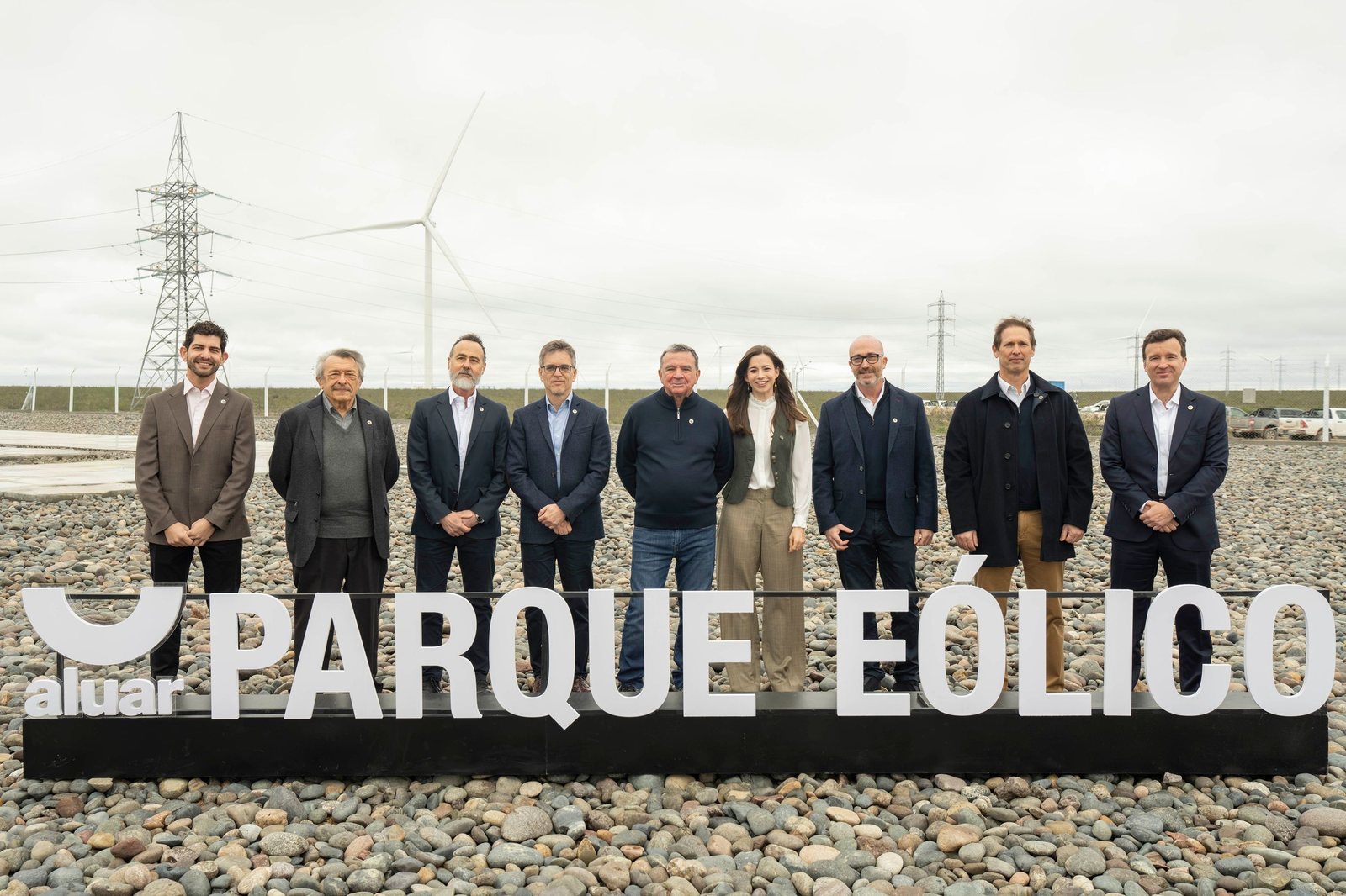 Grupo de directivos frente al cartel de Aluar Parque Eólico, con un aerogenerador y torres de alta tensión detrás